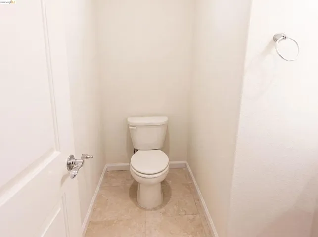 Compact water closet featuring neutral-toned walls, a white toilet, light-colored tile flooring, white baseboards, and a chrome towel ring
