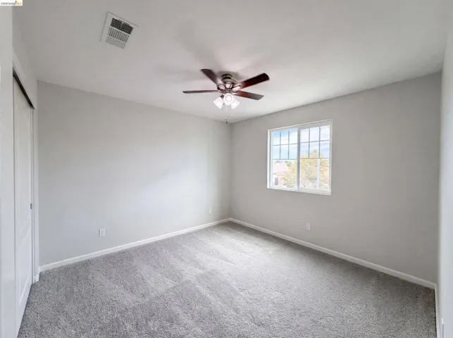 Spacious room featuring light gray walls, gray carpet flooring, a window with gridded panes, a dark-toned ceiling fan with integrated lighting, and a white bi-fold closet door