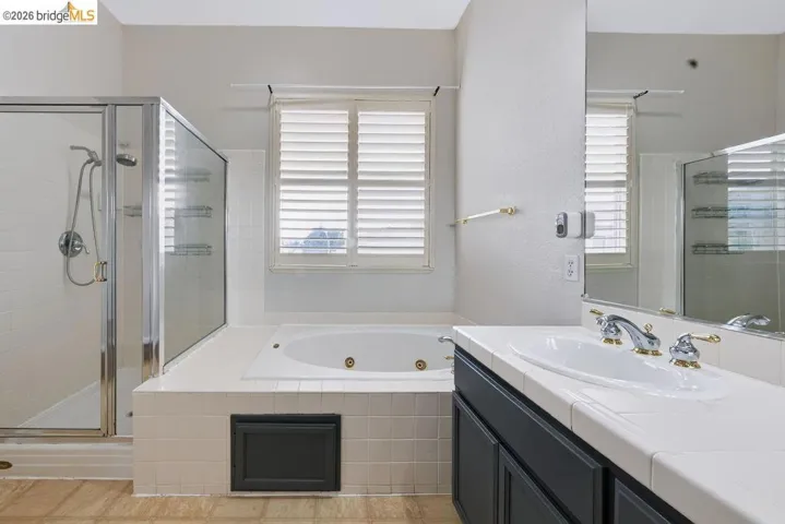 Bathroom featuring a jetted tub with white tile surround, a glass-enclosed shower, dual vanity with white tiled countertop, and a window with plantation shutters