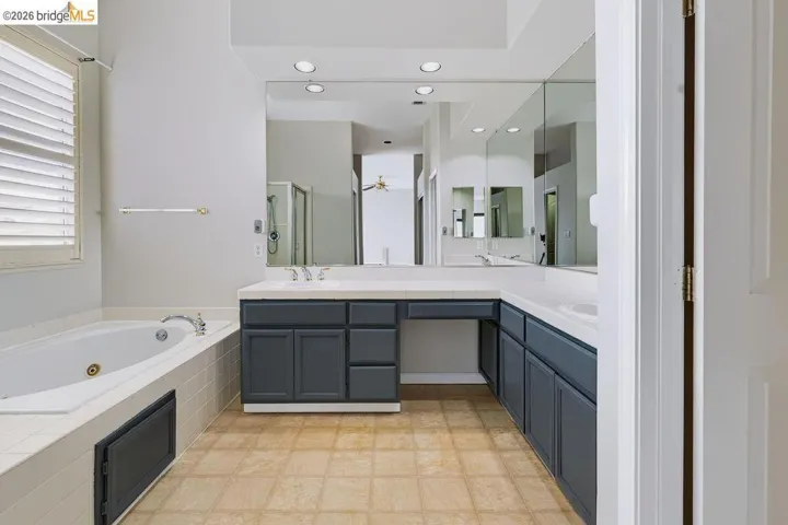 Bathroom featuring a white jetted tub, plantation shuttered window, extensive vanity with dual sinks, dark cabinetry, and full-wall mirrors