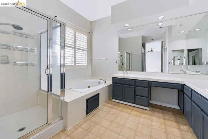 Bathroom featuring a walk-in shower with glass enclosure, a jetted soaking tub, dark grey cabinetry, a vanity with dual sinks, and large wall-mounted mirrors