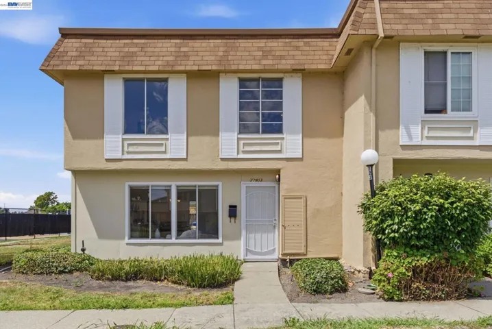 Two-story stucco facade featuring a mansard-style roof with brown shingles, multiple windows with white louvered shutters, a white security screen door, and a globe light fixture