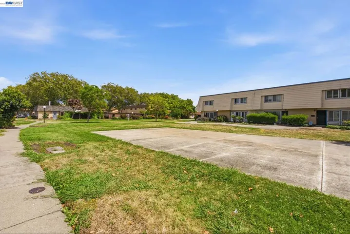 Paved concrete area with surrounding green space, mature trees, and a residential building featuring multiple windows and a dark entry door