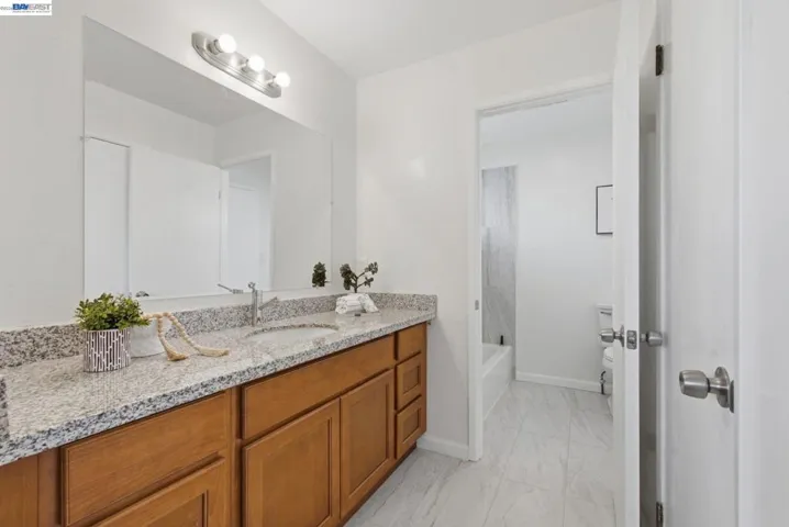 Bathroom featuring a light-toned granite-style double vanity with integrated sinks, wood-finish cabinetry, a large wall-mounted mirror, and a three-bulb vanity light fixture