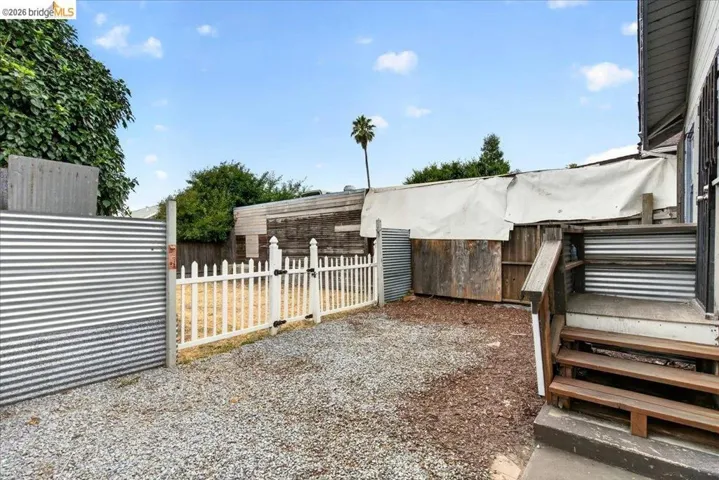 Gravel surface backyard area featuring wood stairs, a white picket fence, and corrugated metal fencing