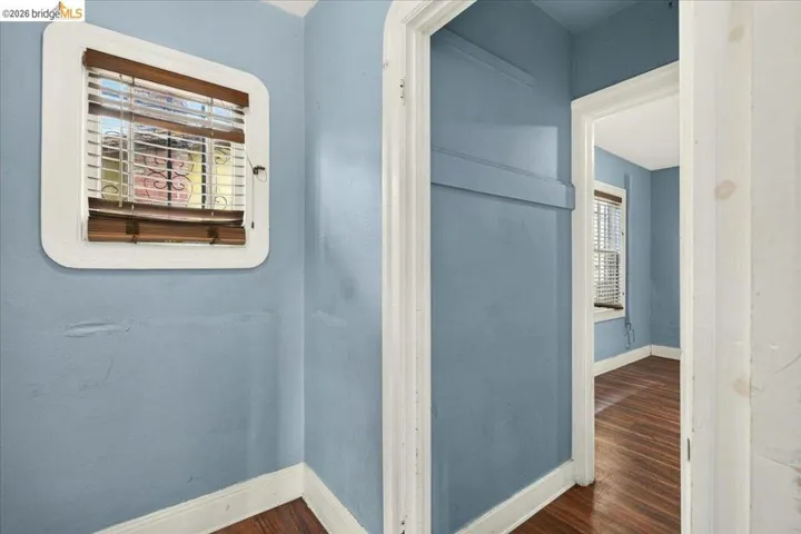 Interior hallway featuring a window with wood-finish blinds, light blue walls, white trim, and wood-finish flooring extending into an adjacent room