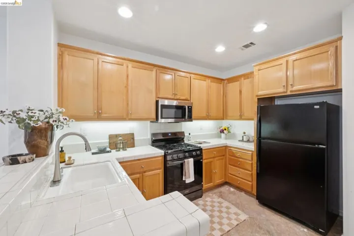 Kitchen featuring light wood cabinetry, white tiled countertops, stainless steel microwave, black gas range, and a black refrigerator