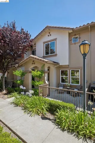 Multi-story stucco exterior featuring a covered entry, tile roof accents, ground-level patio with metal railing, and established landscaping