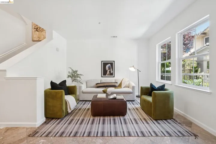 Bright living area with two large windows, white walls, light-toned tile flooring, and staircase with white railing