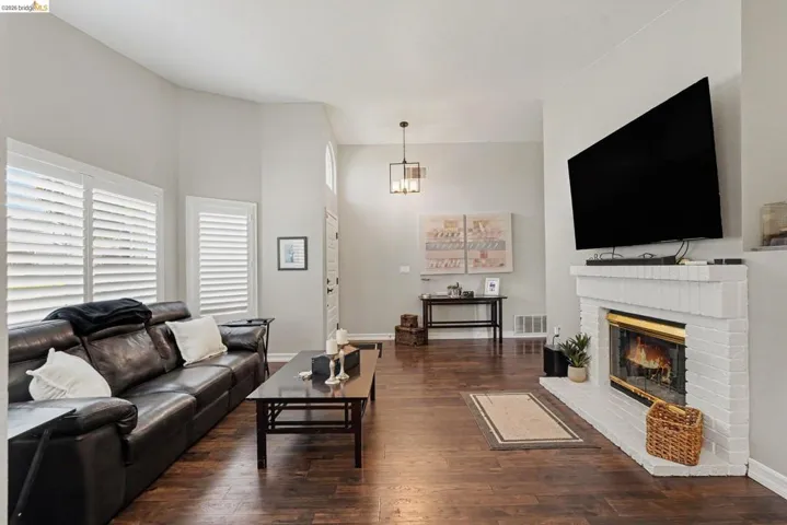Living room featuring wood-finish flooring, a white painted brick fireplace with a gold-framed firebox, white plantation shutters, vaulted ceilings, and a modern ceiling light fixture