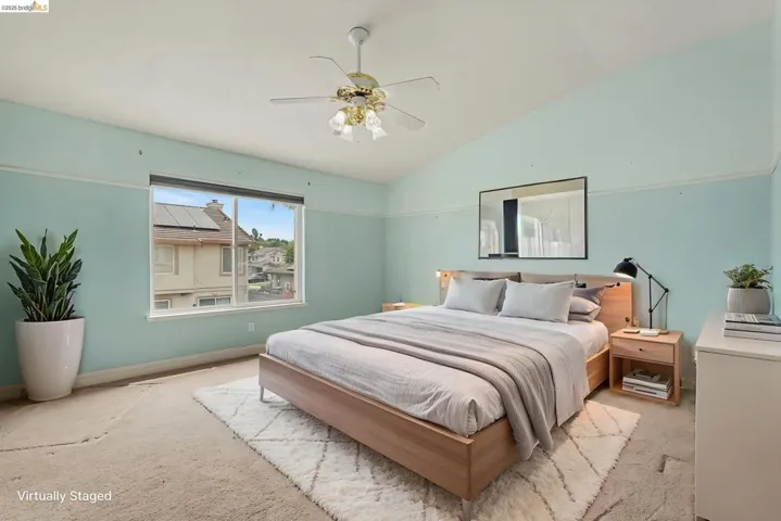 Vaulted ceilings highlight this bedroom, featuring a large window, a ceiling fan with integrated lighting, and light-colored walls with trim detailing