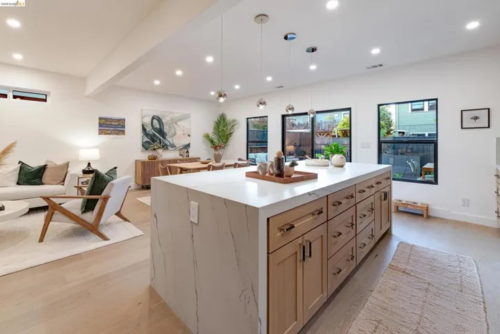 Spacious open-concept living area featuring an oversized kitchen island with a stone-finish countertop, recessed lighting, wood-finish flooring, and multiple large windows with black framing