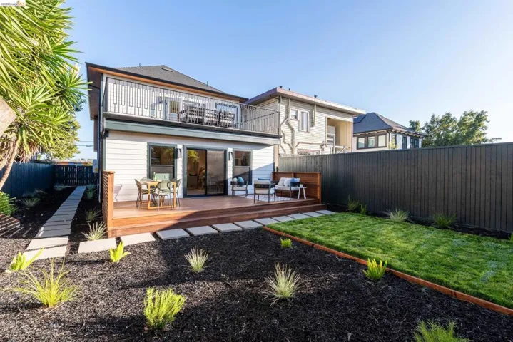 Rear exterior featuring a large wooden deck, sliding glass doors, a second-story balcony with metal railings, a manicured lawn area, and a dark privacy fence