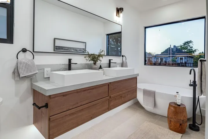 Modern bathroom featuring a floating wood-finish double vanity with rectangular vessel sinks, matte black fixtures, a large framed mirror, and a freestanding soaking tub under a picture window