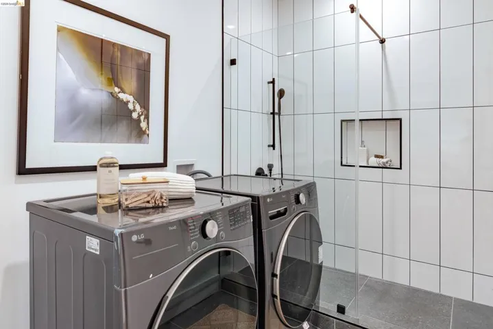 Contemporary laundry room featuring a dark gray washer and dryer set, white vertical subway tile, a glass shower enclosure, and dark gray floor tiling