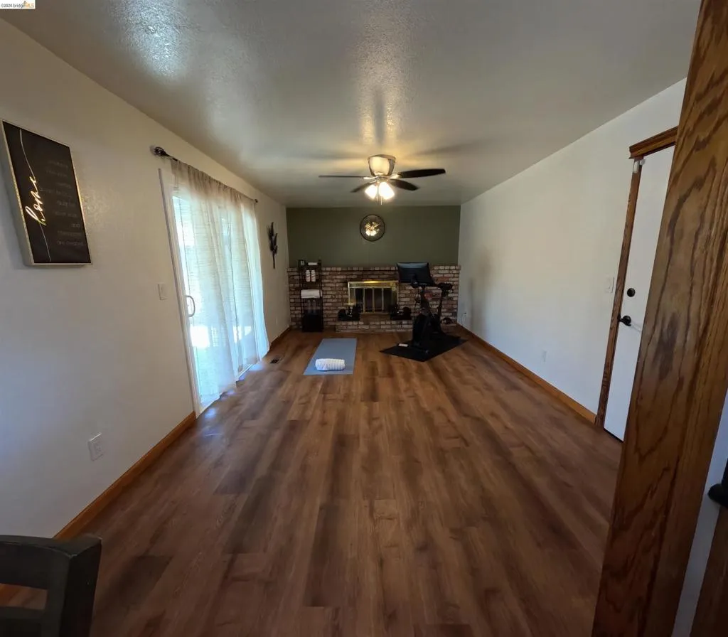 Spacious interior room featuring wood-finish flooring, a brick fireplace with a mantel, and a ceiling fan with integrated lighting