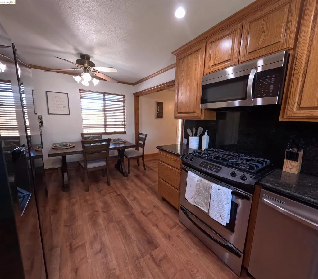 Kitchen and dining area featuring wood-finish flooring, stainless steel appliances, wood cabinetry, dark countertops, and a ceiling fan