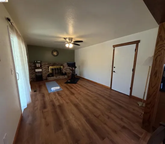 Spacious room featuring wood-finish flooring, a brick fireplace with a brass-toned insert, and a ceiling fan with integrated lighting