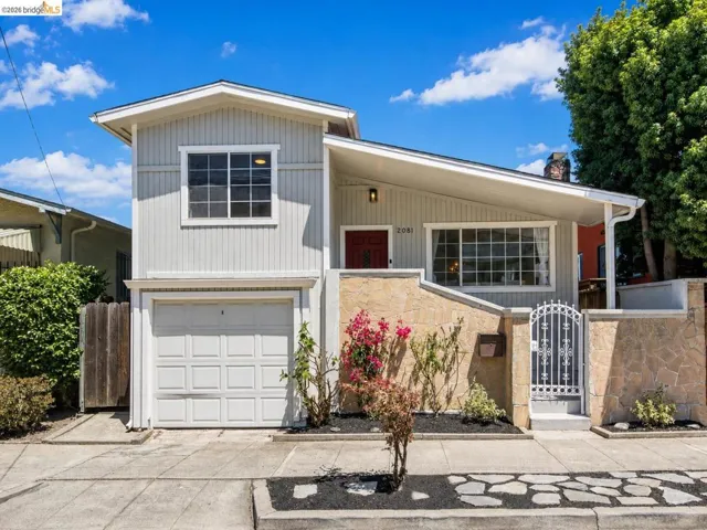 Light gray vertical siding exterior with a red entry door, an integrated garage, a stone facade, a decorative metal pedestrian gate, and a covered front entry