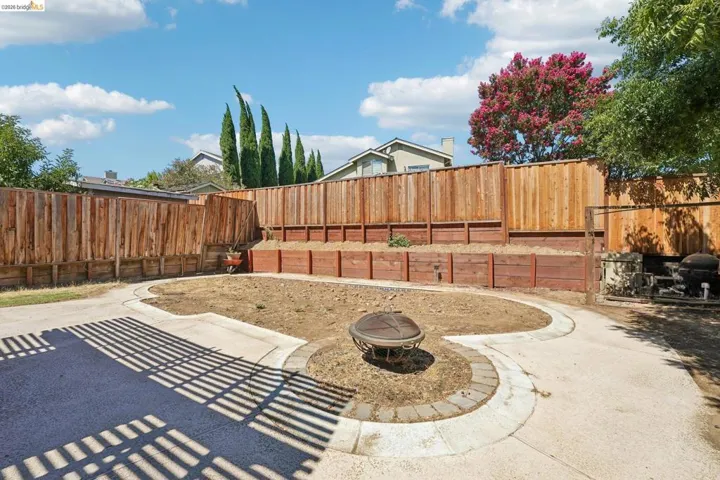 Expansive backyard featuring a circular paver fire pit area, concrete pathways, a tiered wooden retaining wall, and privacy fencing
