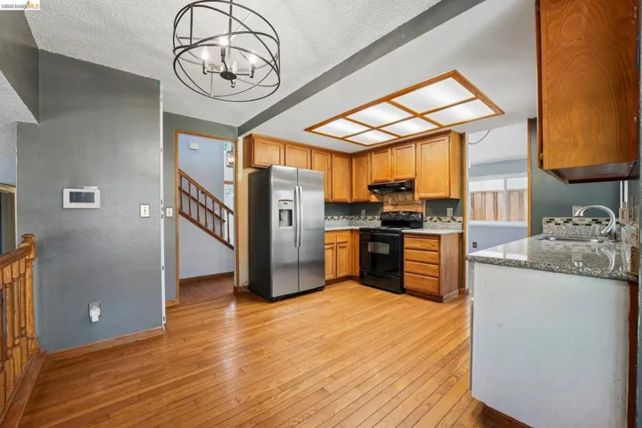 Kitchen featuring natural wood cabinetry, stainless steel refrigerator, black range, granite countertops, and wood-finish flooring