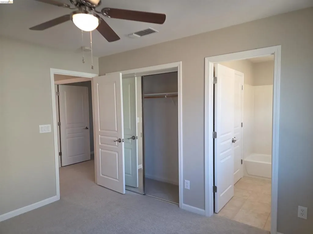 Carpeted room featuring a ceiling fan with integrated lighting, mirrored closet doors, and access to a full bathroom with a tub/shower combination