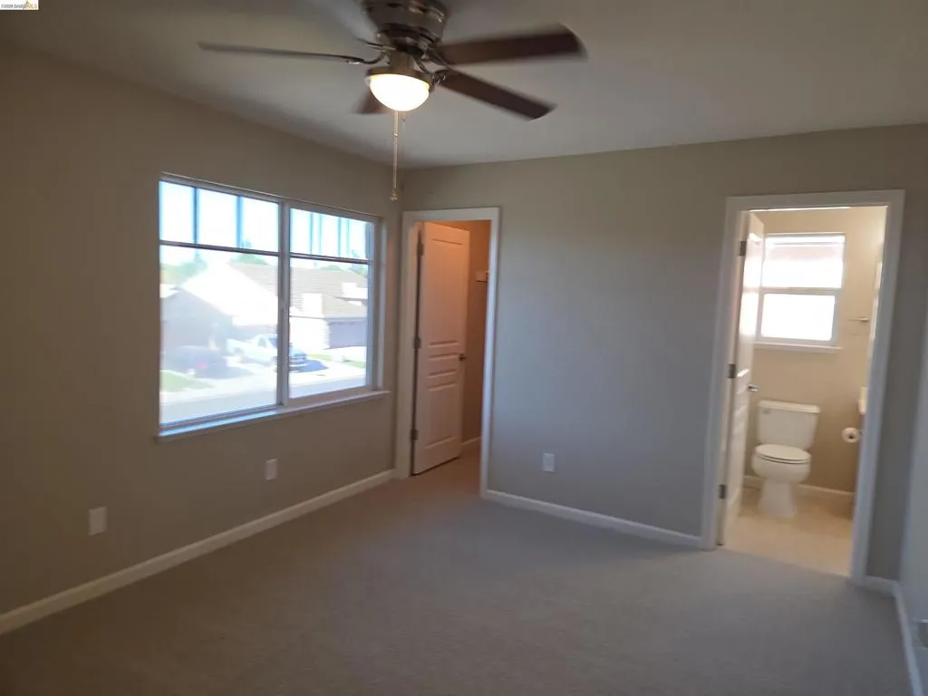 Carpeted room with a ceiling fan light fixture, multiple windows, and a door leading to a bathroom with a window