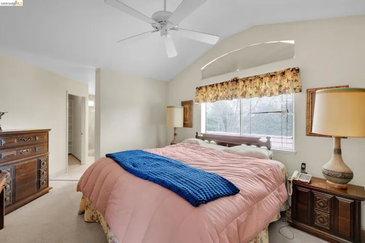 Spacious bedroom featuring a vaulted ceiling with a fan, a large window with blinds, and an arched transom window