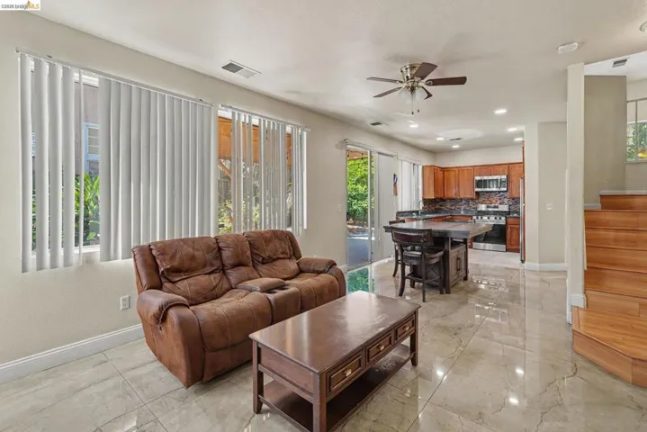 Open-concept living area featuring large windows with vertical blinds, polished stone-finish flooring, a kitchen with wood cabinetry and stainless steel appliances, and a wood-finish staircase