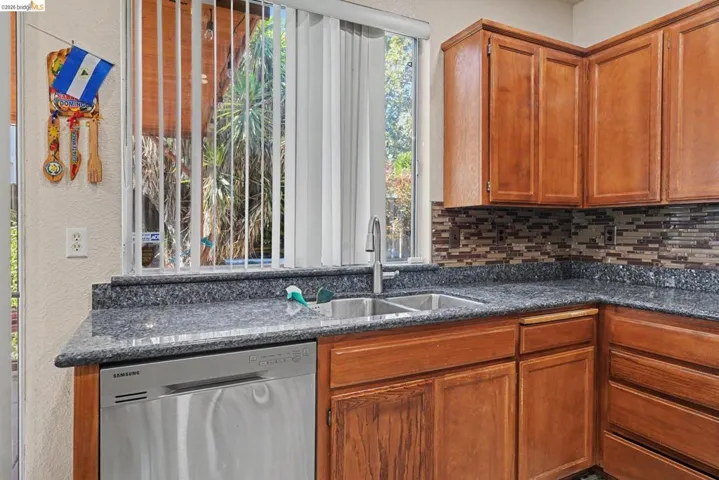 Kitchen area featuring a double basin stainless steel sink, a stainless steel dishwasher, wood cabinetry, a mosaic tile backsplash, and a window with vertical blinds