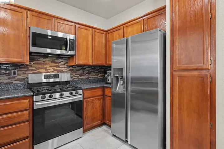 Kitchen featuring wood cabinetry, stainless steel appliances, a multi-tone tile backsplash, and dark speckled countertops