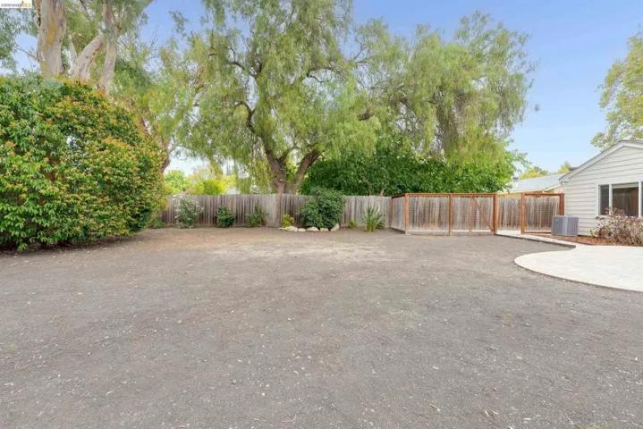 Expansive rear grounds featuring a wood privacy fence, mature trees, a manicured shrub line, and a concrete patio