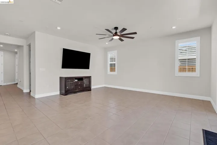 Spacious room featuring tile flooring, neutral wall tones, two windows with plantation shutters, a ceiling fan with integrated lighting, and recessed ceiling lights