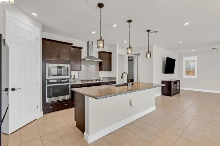 Spacious kitchen featuring an island with a built-in sink, stainless steel appliances, dark wood-finish cabinetry, recessed lighting, and pendant lights