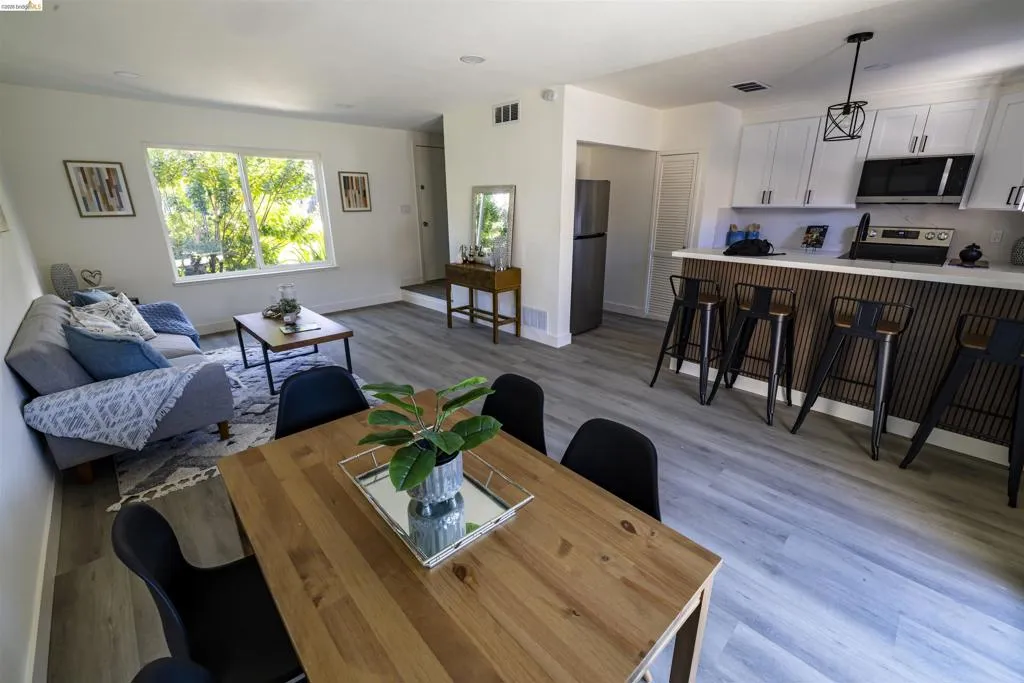Bright living area featuring wood-finish flooring, a contemporary kitchen with white cabinetry, a breakfast bar, and a large window