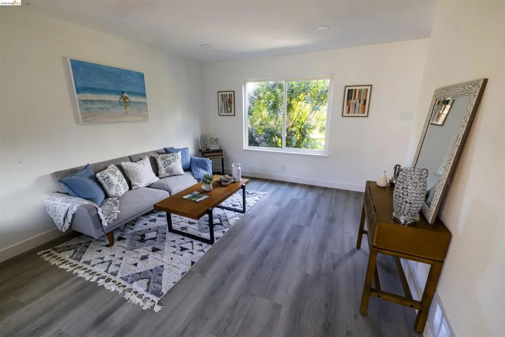 Spacious living area featuring light wood-finish flooring, recessed lighting, white walls, and a large window overlooking lush greenery