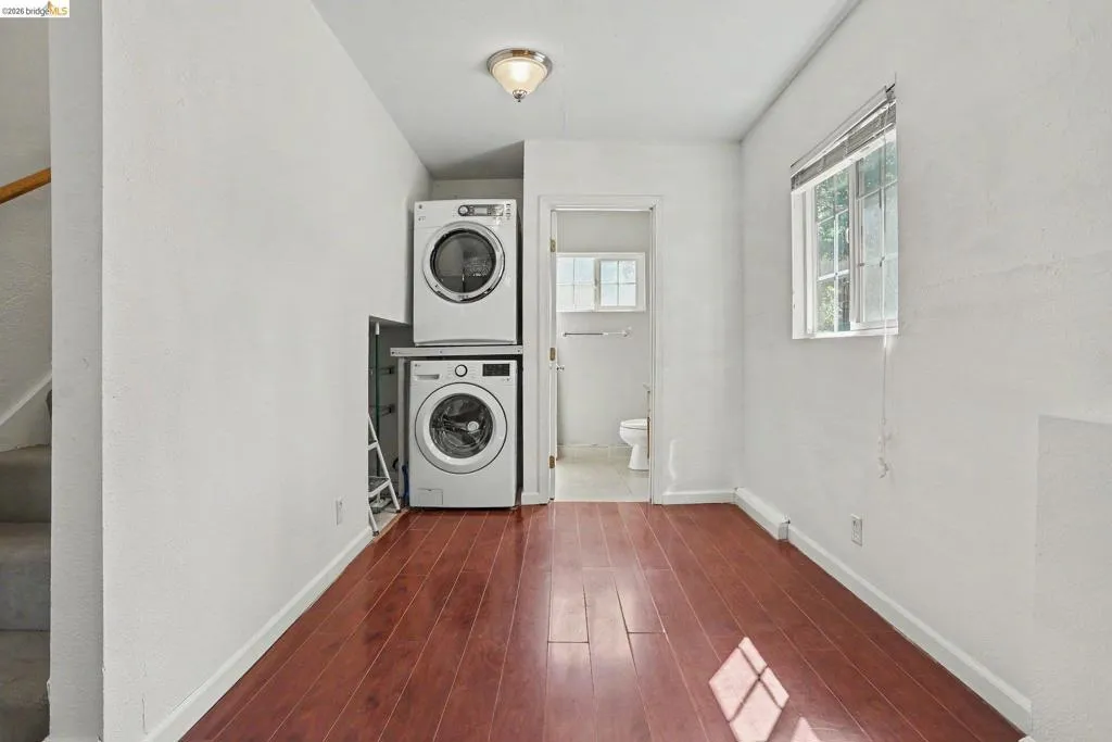 Laundry area featuring a stacked washer and dryer, wood-finish flooring, a window with horizontal blinds, and a ceiling-mounted light fixture