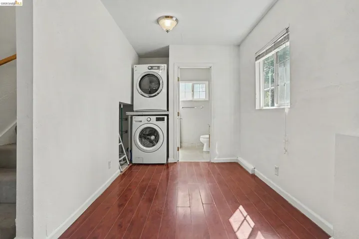 Laundry area featuring a stacked washer and dryer, wood-finish flooring, a window with horizontal blinds, and a ceiling-mounted light fixture