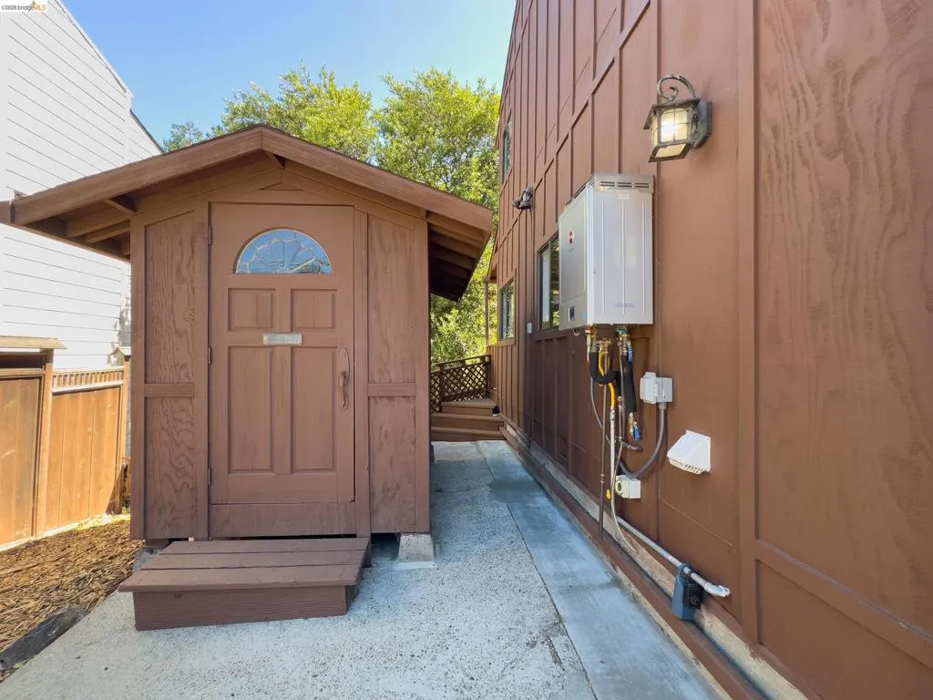Exterior utility space featuring a detached storage shed with a paneled door and transom window, a tankless water heater, and a decorative wall-mounted outdoor light fixture