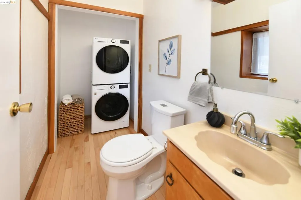 Bathroom featuring a vanity with an integrated sink, a toilet, a stacked washer and dryer, wood-finish flooring, and wood trim accents