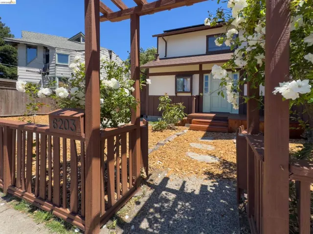 Front entry featuring a wooden pergola with climbing white roses, a decorative wooden fence with house numbers, and a light blue front door