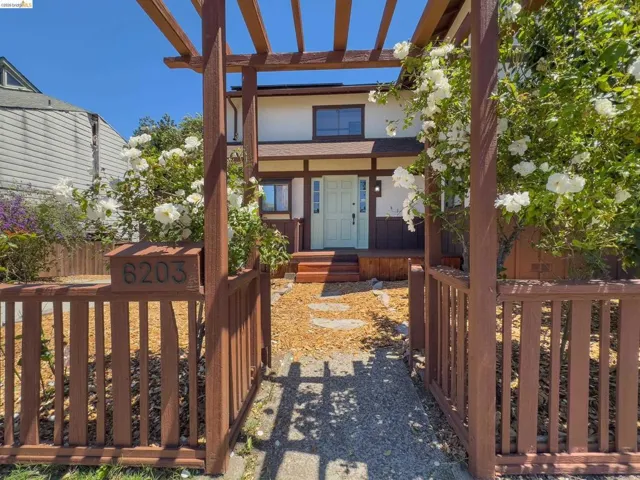 Charming entryway featuring a wooden pergola with climbing white roses, a decorative stone pathway, a wooden picket fence, and a welcoming light blue front door