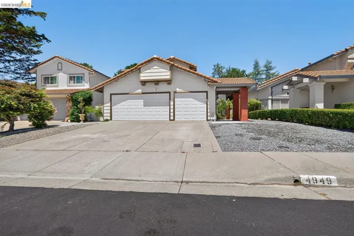 Stucco exterior with a tile roof, featuring a two-car garage with white paneled doors, a paved driveway, and a gravel front yard
