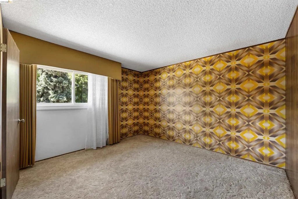 Room featuring a patterned accent wall, light-colored carpet flooring, a window with exterior tree views, and an overhead textured ceiling