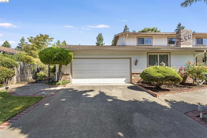 Two-car garage with a sectional door, concrete driveway, brick perimeter landscaping, and a wrought iron gate with an entry path