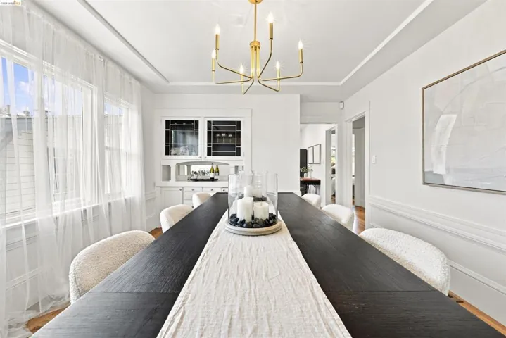 Dining area featuring a contemporary gold chandelier, built-in cabinetry with glass fronts, white wainscoting, and large windows with sheer coverings