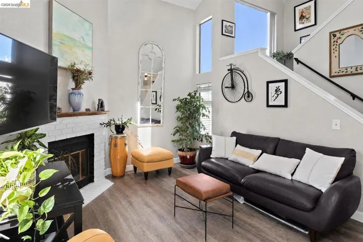 Living area featuring high ceilings, wood-finish flooring, a white brick fireplace with a wood mantel, large windows, and an open staircase with a black handrail