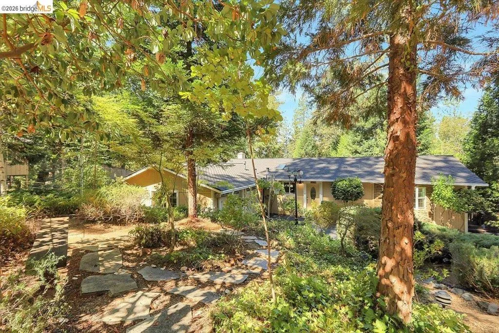 Mature landscaping with established trees frames a single-story residence featuring a neutral-toned exterior, dark shingle roof, and a stone-paver walkway