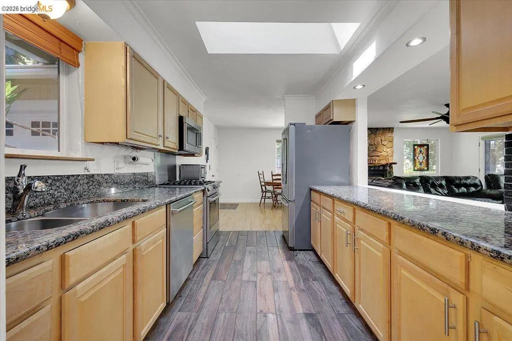 Kitchen featuring wood-finish flooring, light wood cabinetry, stainless steel appliances, granite countertops, and a skylight