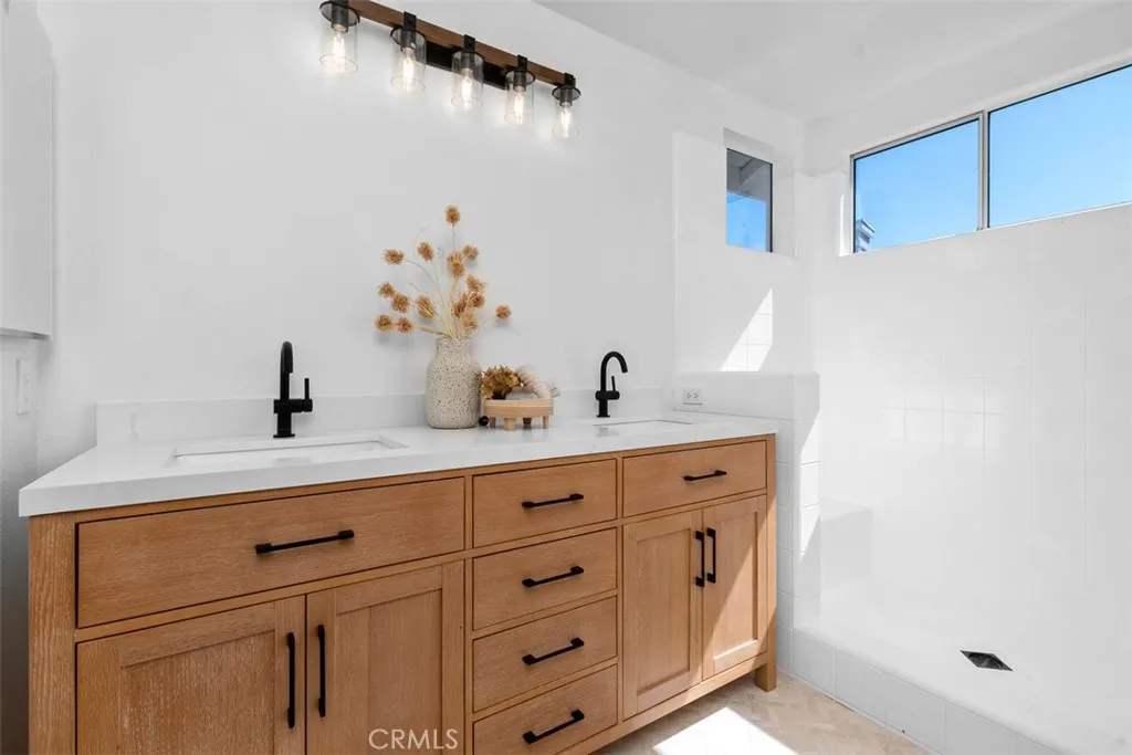 Primary Bathroom: Double-sink vanity with warm wood cabinetry, black fixtures, and a bright white countertop sits beside a tiled walk-in shower with natural light from clerestory windows.